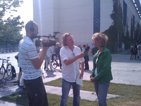 Ein Journalist und ein Kameramann interviewen eine mit grünem Oberteil bekleidete Frau.