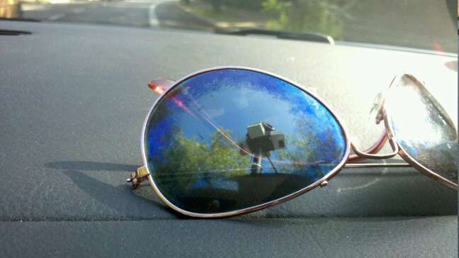 Sonnenbrille auf der Ablage eines Autos, in der sich ein die Frontscheibe, Bäume und ein auf dem Dach befindlicher Kamera-Aufbau spiegeln
